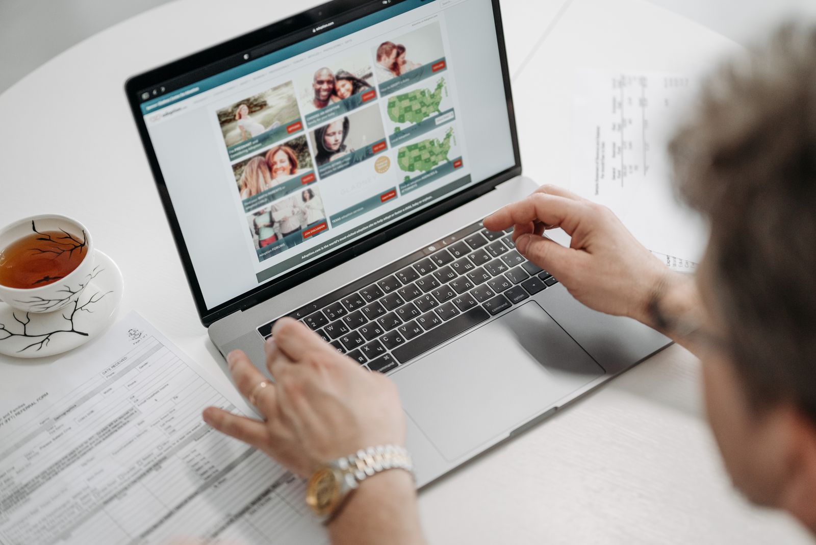 A person browsing a laptop beside papers and a cup of tea